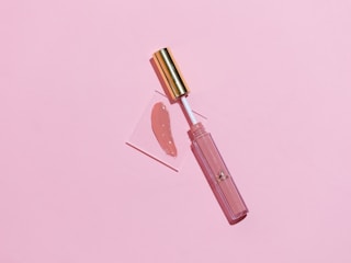 A makeup lip gloss with a gold cap lies on a soft pink background. The gloss is slightly opened and the applicator is placed on a small transparent square with a swatch of the lip gloss color beside it.