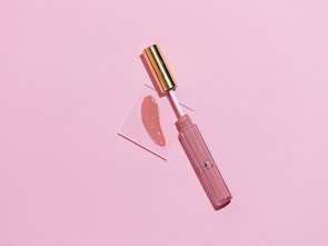 A makeup lip gloss with a gold cap lies on a soft pink background. The gloss is slightly opened and the applicator is placed on a small transparent square with a swatch of the lip gloss color beside it.