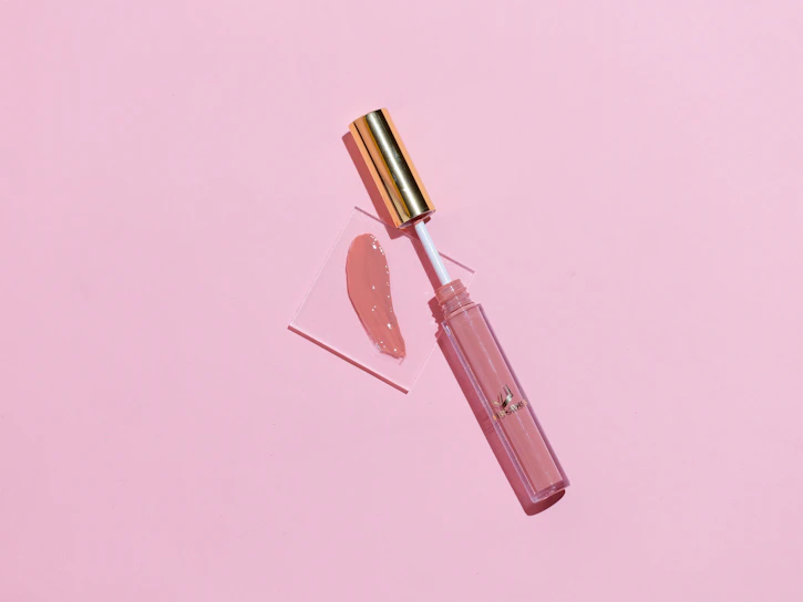Close-up of a glossy lip oil dripping softly on a pastel pink background with subtle light reflections.