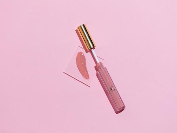 A makeup lip gloss with a gold cap lies on a soft pink background. The gloss is slightly opened and the applicator is placed on a small transparent square with a swatch of the lip gloss color beside it.