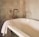 A vintage-style bathroom featuring a freestanding bathtub with a white towel draped over the edge. The wall is adorned with light-colored tiles and marble accents. The bathtub faucet and showerhead have a brass finish, adding a touch of elegance.