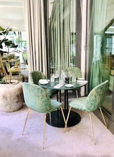 A modern outdoor dining setup with a round black table surrounded by four green upholstered chairs with gold legs. The table is elegantly set with white plates, crystal glasses, cutlery, and a black pepper mill. Tall curtains and glass windows in the background add a sophisticated touch, and a large potted plant is visible to the side.