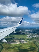 Aerial view of a landscape featuring a partially visible airplane wing against a bright blue sky with scattered white clouds. Below lies a sprawling green terrain interspersed with both developed areas and patches of open ground.