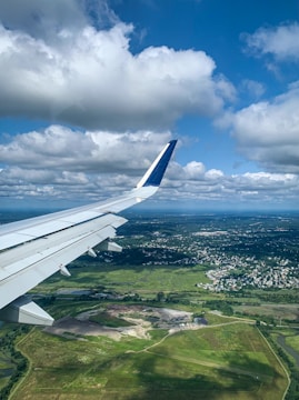 Aerial view of a landscape featuring a partially visible airplane wing against a bright blue sky with scattered white clouds. Below lies a sprawling green terrain interspersed with both developed areas and patches of open ground.