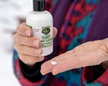A person holds a bottle of CBD body lotion with the label '1000 MG CBD, Eden's CBD, 100% Pure CBD' and dispenses a small amount onto their palm. The person is wearing a colorful, patterned fabric in the background.