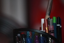 A mesh holder filled with a variety of writing utensils, including pens, markers, and possibly a paintbrush. The focus is on the holder, which contains a mix of colorful items with a blurred red background.