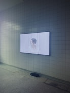 A wall-mounted screen displays an image of a woman with dark hair tied back. The screen is set against a white tiled wall in a room with dim lighting. The floor appears to be made of a light-colored stone or concrete material. A black soundbar or speaker is positioned just below the screen.