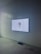 A wall-mounted screen displays an image of a woman with dark hair tied back. The screen is set against a white tiled wall in a room with dim lighting. The floor appears to be made of a light-colored stone or concrete material. A black soundbar or speaker is positioned just below the screen.