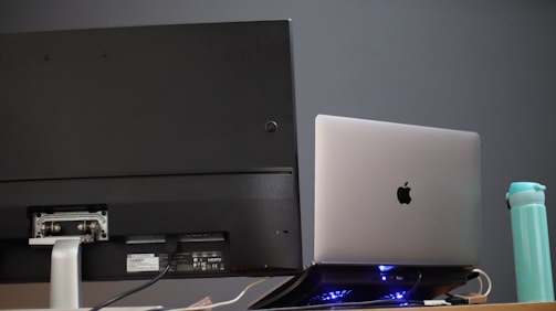 A desktop monitor and a laptop placed side by side on a desk. The laptop is elevated with a stand and has a glowing base. A reusable water bottle is positioned nearby. The monitor has various ports and labels visible on the back.