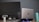 A desktop monitor and a laptop placed side by side on a desk. The laptop is elevated with a stand and has a glowing base. A reusable water bottle is positioned nearby. The monitor has various ports and labels visible on the back.