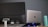 A desktop monitor and a laptop placed side by side on a desk. The laptop is elevated with a stand and has a glowing base. A reusable water bottle is positioned nearby. The monitor has various ports and labels visible on the back.