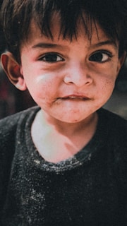 A young child with short, dark hair and large, expressive eyes gazes directly at the camera. The child's face is slightly dirty, adding a playful or adventurous character to the expression. Wearing a dark shirt, the overall tone of the image is muted and soft.