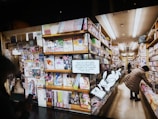 A bookstore aisle filled with shelves stocked with brightly colored manga volumes. A person browses the shelves on the right side, while another person is partially visible holding a phone. A cartoon rabbit character holding a book with a QR code invites readers to access free manga online.