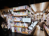 A bookstore aisle filled with shelves stocked with brightly colored manga volumes. A person browses the shelves on the right side, while another person is partially visible holding a phone. A cartoon rabbit character holding a book with a QR code invites readers to access free manga online.