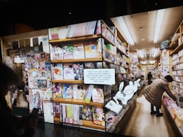 A bookstore aisle filled with shelves stocked with brightly colored manga volumes. A person browses the shelves on the right side, while another person is partially visible holding a phone. A cartoon rabbit character holding a book with a QR code invites readers to access free manga online.