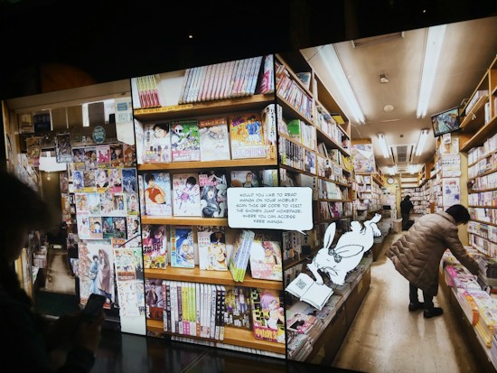 A bookstore aisle filled with shelves stocked with brightly colored manga volumes. A person browses the shelves on the right side, while another person is partially visible holding a phone. A cartoon rabbit character holding a book with a QR code invites readers to access free manga online.