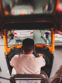 A person is driving a yellow and black auto rickshaw in traffic, surrounded by several cars visible through the windshield. The image is taken from behind the driver, showcasing the view of the road ahead.