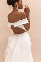 A woman in an elegant, off-the-shoulder white dress with a large bow at the back stands against a neutral background. She has her hair styled in an updo and is wearing large, ornate earrings. Her hands are gently clasped together, conveying a sense of calm and grace.