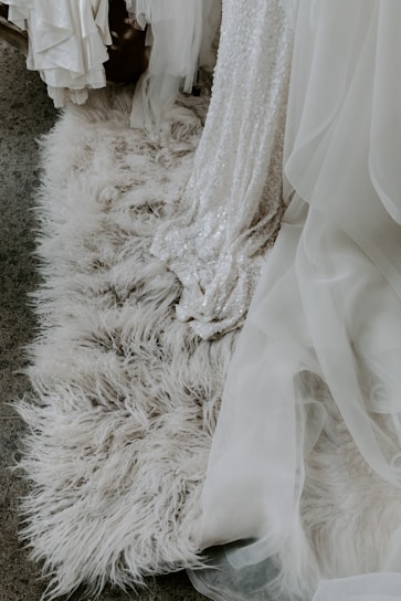 An intricate white dress with sequins draped elegantly over a plush, white, fluffy rug. The scene suggests a bridal setting with layers of tulle and fabric visible.