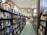 A bookstore aisle lined with shelves filled with a variety of books. The store is well-lit with high ceilings and features arched windows. Toward the back, a small cafe is visible with tables and chairs.