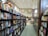 A bookstore aisle lined with shelves filled with a variety of books. The store is well-lit with high ceilings and features arched windows. Toward the back, a small cafe is visible with tables and chairs.
