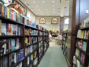 A bookstore aisle lined with shelves filled with a variety of books. The store is well-lit with high ceilings and features arched windows. Toward the back, a small cafe is visible with tables and chairs.