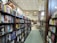 A bookstore aisle lined with shelves filled with a variety of books. The store is well-lit with high ceilings and features arched windows. Toward the back, a small cafe is visible with tables and chairs.