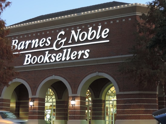 A Barnes & Noble bookstore with prominent signage on the exterior of a brick building. The front of the store features large arched windows and a well-lit interior. There are trees partially visible on the edges of the image.