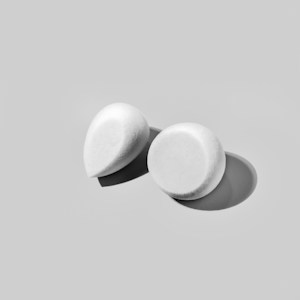 Two white cosmetic sponges, one with a teardrop shape and the other round, are positioned on a smooth, light gray background. Soft shadows are cast beneath them, indicating a light source from above.
