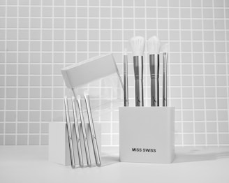 A collection of makeup brushes is neatly arranged in a white container against a tiled wall background. The container has a minimalist design and is labeled 'Miss Swiss'. The brushes have sleek, metallic handles with soft, white bristles. Several are standing upright in the container, while others are leaning against it.