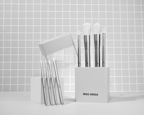 A collection of makeup brushes is neatly arranged in a white container against a tiled wall background. The container has a minimalist design and is labeled 'Miss Swiss'. The brushes have sleek, metallic handles with soft, white bristles. Several are standing upright in the container, while others are leaning against it.