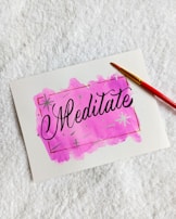 A white piece of paper lying on a textured white surface. The paper features a hand-painted pink watercolor background with the word 'Meditate' written in elegant black script in the center. A red-handled paintbrush with a yellow tip is placed diagonally across the top right corner of the paper.