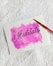 A white piece of paper lying on a textured white surface. The paper features a hand-painted pink watercolor background with the word 'Meditate' written in elegant black script in the center. A red-handled paintbrush with a yellow tip is placed diagonally across the top right corner of the paper.