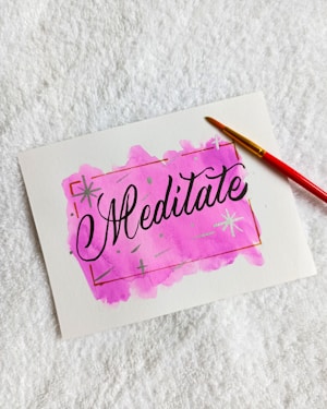 A white piece of paper lying on a textured white surface. The paper features a hand-painted pink watercolor background with the word 'Meditate' written in elegant black script in the center. A red-handled paintbrush with a yellow tip is placed diagonally across the top right corner of the paper.