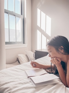 A person with long hair is lying on a bed, writing in a notebook with a pen. Sunlight is streaming through a window, creating patterns of light and shadow on the walls. The atmosphere appears calm and peaceful.