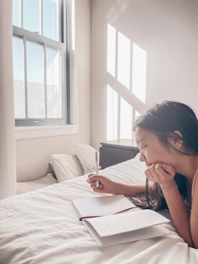 A serene woman writing in a notebook by a window with sunlight gently coming in, symbolizing reflection and faith.