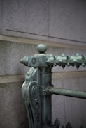 An ornate metal railing with decorative details, featuring a spherical finial on top and a series of pointed spikes along the edge. The surface has a weathered green patina, suggesting it is made of aged bronze or copper. The background wall is made of large, light gray stone blocks.