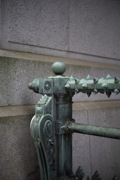 An ornate metal railing with decorative details, featuring a spherical finial on top and a series of pointed spikes along the edge. The surface has a weathered green patina, suggesting it is made of aged bronze or copper. The background wall is made of large, light gray stone blocks.