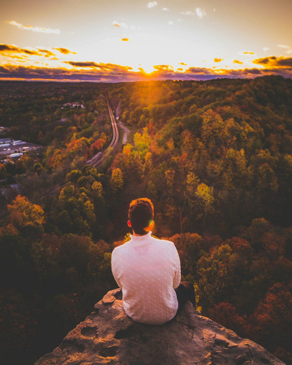 Mann in weißem Rundhals-T-Shirt sitzt auf Felsen und schaut bei Sonnenuntergang auf die Berge