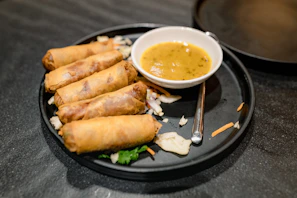 Fresh spring rolls crisply fried and served alongside a spicy orange-hued sauce, styled on a black slate.