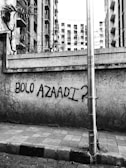 A concrete wall, covered with barbed wire on top, has graffiti that reads 'BOLO AZAADI?' A tall metal pole stands in front of the wall, with buildings in the background.