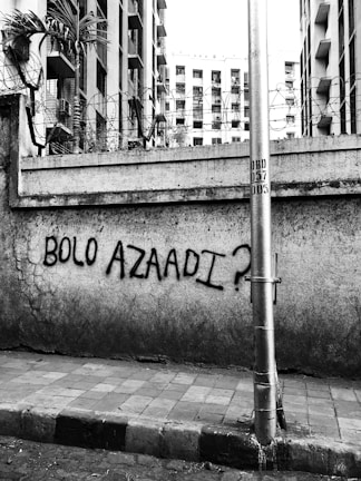 A concrete wall, covered with barbed wire on top, has graffiti that reads 'BOLO AZAADI?' A tall metal pole stands in front of the wall, with buildings in the background.