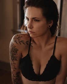 A person with tattoos is wearing a black lace bralette. They have a contemplative expression and a nose ring and ear gauges. The lighting is warm and soft, adding a serene and introspective mood to the image.