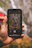 A hand is holding a smartphone displaying a photograph of four individuals sitting in front of a waterfall. The background shows blurred natural scenery, likely the waterfall and surrounding greenery.