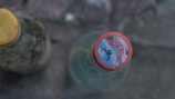 Two glass bottles are captured from a top-down perspective. The foreground bottle has a colorful cap with an abstract or logo design, which includes red tones and some text or numbers. The background bottle has a yellow cap and appears blurred, suggesting the use of shallow depth of field.