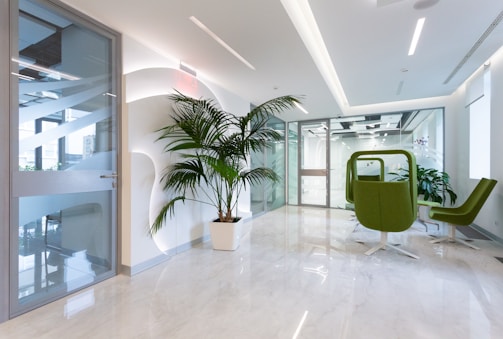 A modern office space with a bright, clean aesthetic. The area features a large potted plant against a white wall with geometric designs. The floor is made of polished, light-colored marble. There are two green swivel chairs and a small table near a glass door. Large glass partitions separate the space, allowing natural light to illuminate the area.