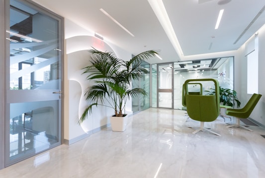 A modern office space with a bright, clean aesthetic. The area features a large potted plant against a white wall with geometric designs. The floor is made of polished, light-colored marble. There are two green swivel chairs and a small table near a glass door. Large glass partitions separate the space, allowing natural light to illuminate the area.