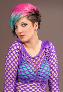 A person with a bold hairstyle featuring pink and blue colors. They are wearing dramatic makeup with black eyeliner and have facial piercings. The outfit includes a purple mesh top over a turquoise undergarment, adding an edgy and expressive style.