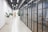 A modern, well-lit office corridor with white brick walls on one side and a glass partition with black frames on the other. The floor is light grey and polished, extending towards a wooden door at the far end. There are several large white planters with green plants along the corridor. Overhead, fluorescent lights and black-colored piping are visible.