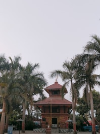 A traditional temple structure with a red brick facade and tiered roof is surrounded by tall palm trees. The sky above is clear, creating a serene and calm atmosphere.